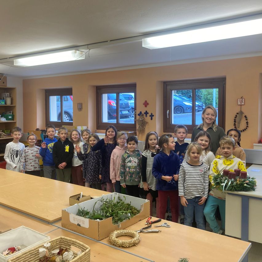 Eine Gruppe von Kindern und Erwachsenen steht in einem Klassenzimmer. Einige tragen Jacken und einige haben Blumen. Es gibt Schreibtische, Kisten und einen Kranz.