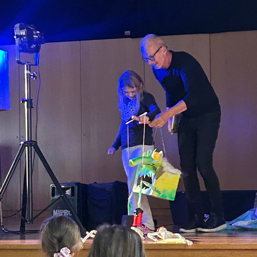 Ein älterer Mann mit Brille steht auf einer Bühne und hält eine Puppe, während ein junges Mädchen neben ihm steht. Auf der linken Seite der Bühne befindet sich ein Stativ mit einer Kamera.