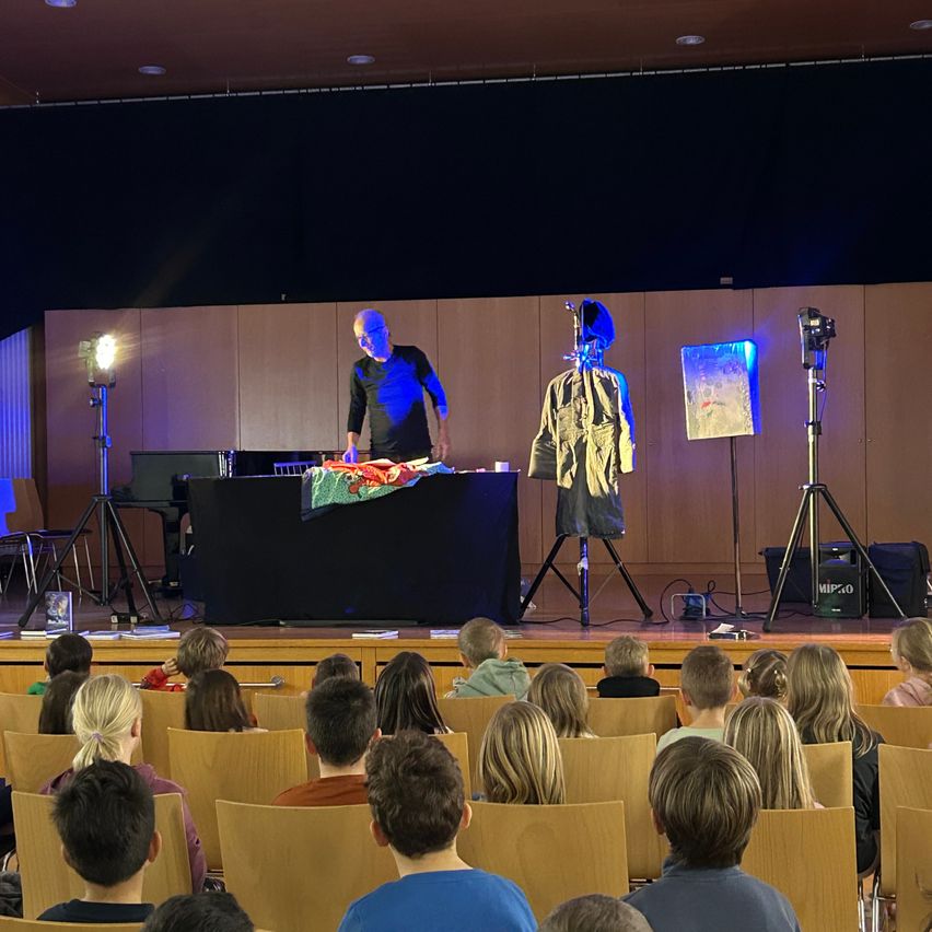 Ein älterer Mann steht auf der Bühne und führt vor einer Gruppe von Kindern einen Zaubertrick vor. Die Bühne ist mit Beleuchtung und Hintergrund ausgestattet.