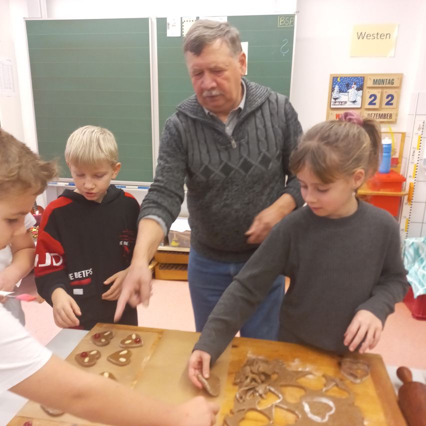 Ein älterer Mann und drei Kinder sind in einem Klassenzimmer und backen Pfefferkuchen. Sie stehen um einen Tisch mit Keksen und Ausstechern.