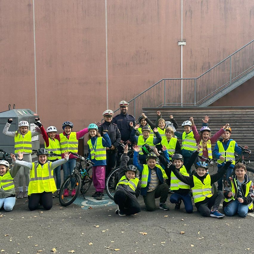 Eine Gruppe von Kindern in leuchtend gelben Warnwesten und Helmen posiert für ein Foto mit Fahrrädern im Freien, mit einem Polizeibeamten und einer Treppe im Hintergrund.