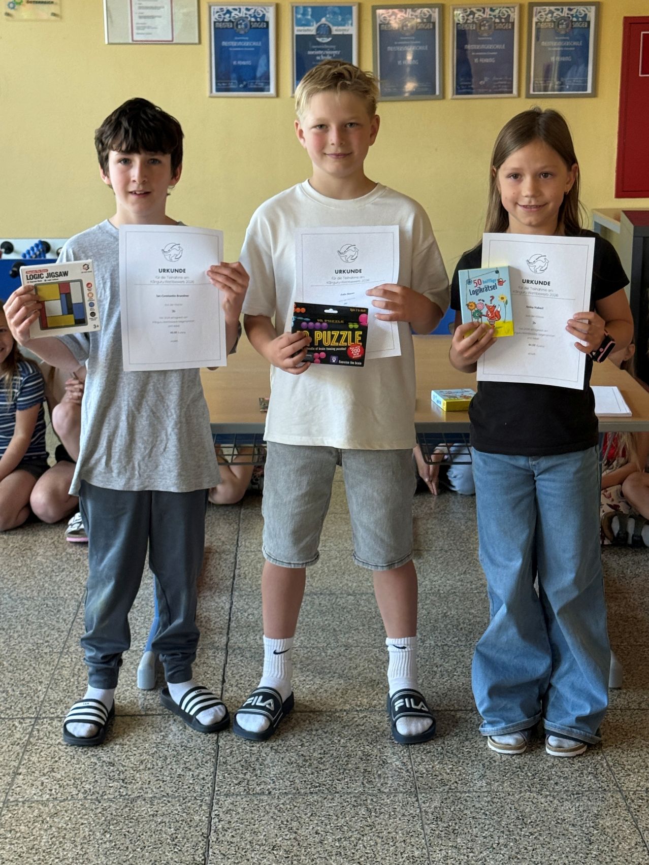 Drei Kinder, zwei Jungen und ein Mädchen, stehen und halten Urkunden und Bücher. Der Junge links hält ein Buch mit dem Titel 'Logic Jigsaw'. Der mittlere Junge hält eine Rätselbox. Das Mädchen rechts hält eine Urkunde.