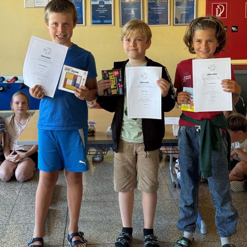 Drei Kinder stehen nebeneinander und halten Urkunden und Preise in der Hand. Der Junge auf der linken Seite trägt ein blaues T-Shirt, der in der Mitte ein grünes und der auf der rechten Seite ein rotes. Sie lächeln. Hinter ihnen sitzen andere Kinder auf dem Boden.