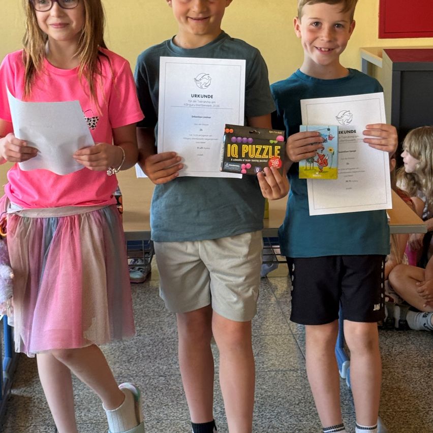 Drei Kinder stehen lächelnd nebeneinander und halten Urkunden in den Händen. Das Mädchen links trägt ein rosa T-Shirt und einen bunten Rock, während die beiden Jungen blaue Shirts und Shorts tragen. Sie halten auch eine Schachtel IQ-Puzzle.