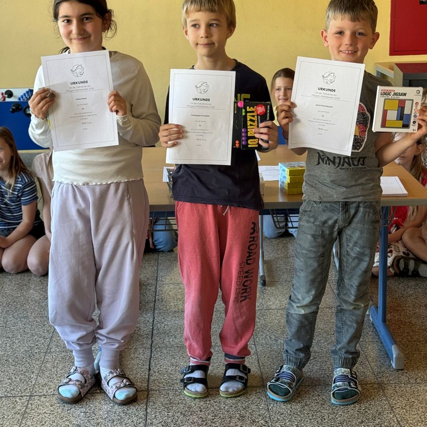 Drei Kinder stehen und halten Urkunden. Ein Mädchen in Weiß, ein Junge in Rot und ein Junge in Grau. Sie alle lächeln und posieren für ein Foto. Dahinter steht ein Tisch mit Büchern und anderen Gegenständen darauf.