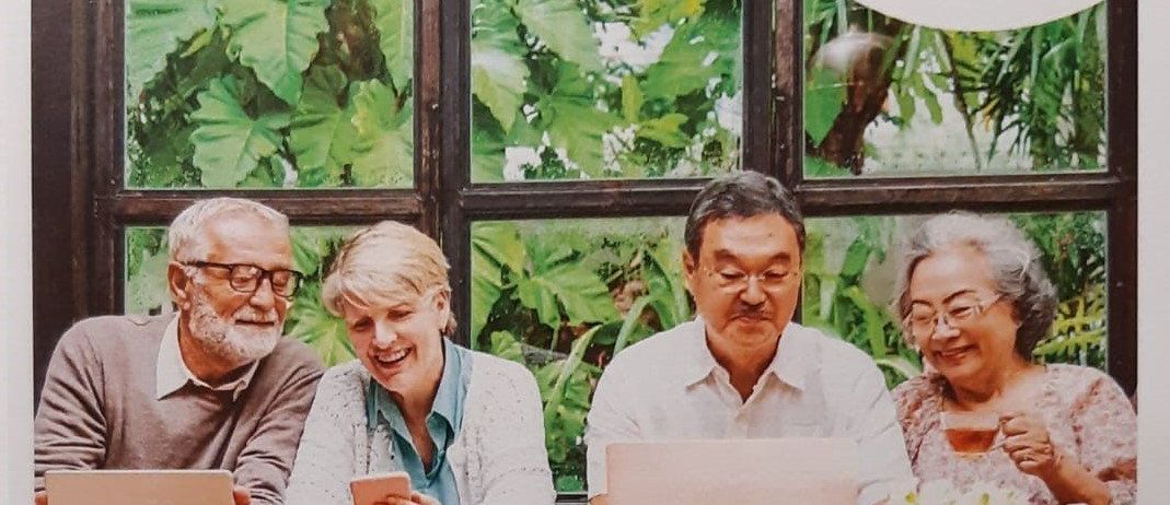 Vier ältere Erwachsene sitzen um einen Tisch herum und nutzen Geräte. Sie lächeln und scheinen engagiert zu sein. Hinter ihnen ein Fenster mit Blick auf tropische Pflanzen.