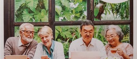 Four elderly people are seated at a table with laptops, smiling and looking at their screens. The setting is outdoors, with large windows behind them showing lush green foliage.