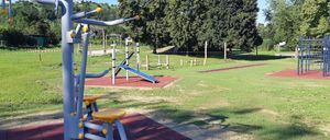 Outdoor-Fitnessgeräte in einem Park mit grünem Rasen und Bäumen. Die Geräte umfassen eine blaue Rutsche und verschiedene Trainingsmaschinen.