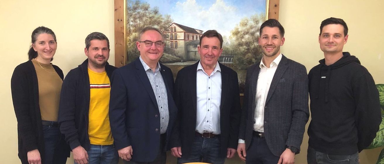 Fünf Männer stehen lächelnd vor einem Gemälde. Sie tragen formelle Kleidung. Der Mann auf der linken Seite trägt ein gelbes Hemd. Hinter ihnen stehen zwei Holzstühle.