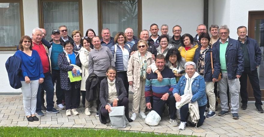Eine Gruppe von Menschen, meist älterer, steht vor einem Gebäude und posiert für ein Foto. Einige tragen eine Brille, und einige halten Bücher. Der Boden ist gepflastert.