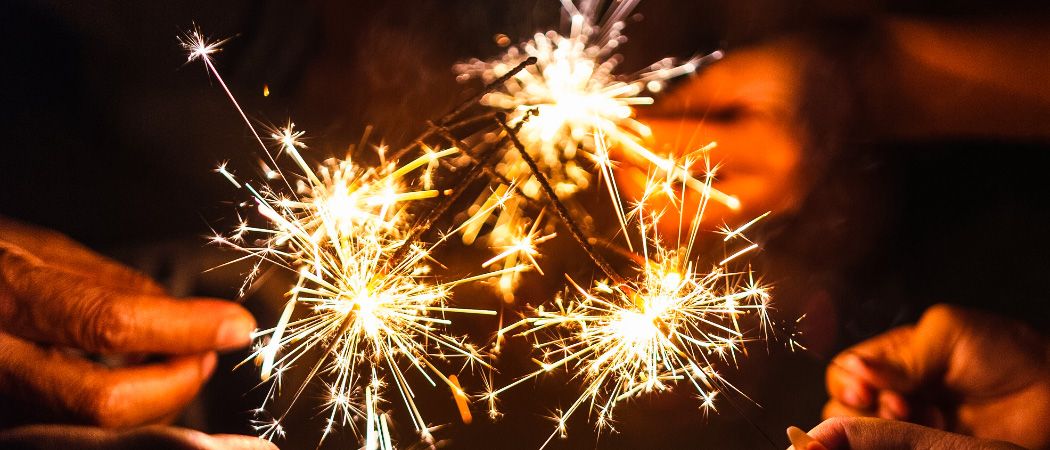 Feuerwerkskörper sind entzündet und beleuchten Hände und Gesichter. Die Funken fliegen nach oben in einem festlichen Display. Eine Person ist im Hintergrund teilweise verschwommen.
