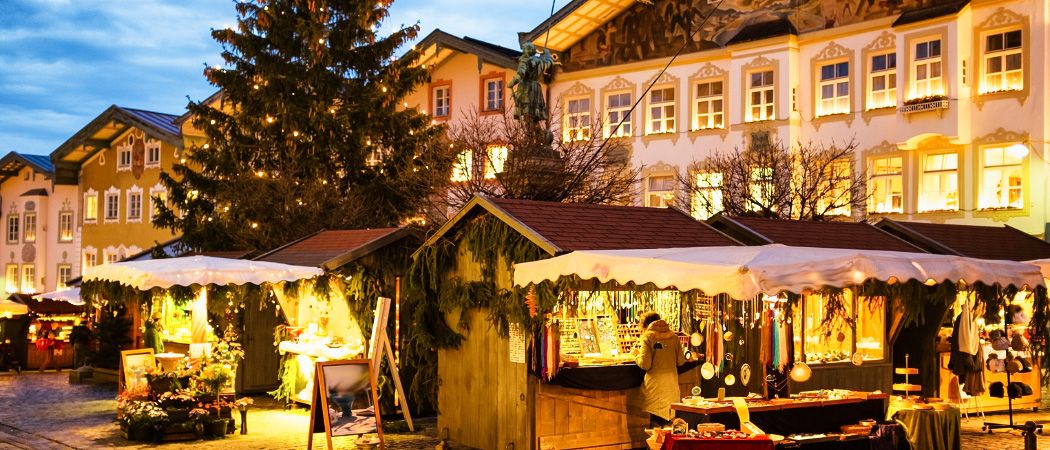 Nächtliche Szene eines Weihnachtsmarktes auf einem Stadtplatz mit kleinen Holzhütten, einem beleuchteten Weihnachtsbaum und einem Gebäude mit verzierten Fenstern.