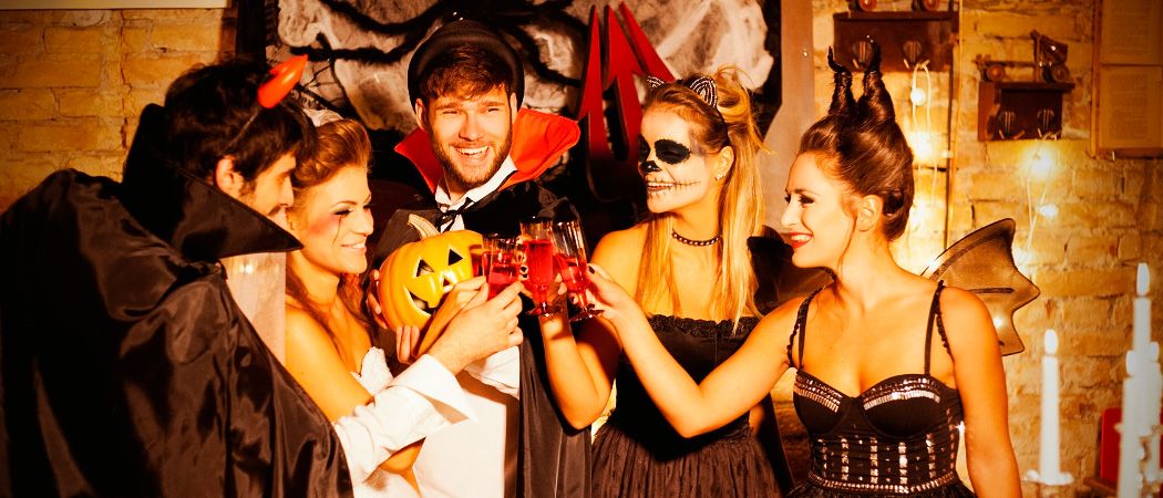 Gruppe von Menschen in Halloween-Kostümen, sie lächeln und stoßen mit Gläsern an, ein Mann in Dracula-Kostüm hält eine Kürbis in der Mitte.