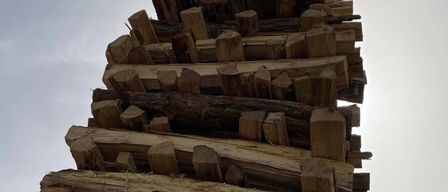 A stack of wood logs is arranged for a bonfire, with a cross on top. The date April 4, 2026 is written on the left. The text reads: 'For a rich bonfire and toast, your FF Deutsch Schützen.' The sky is cloudy.