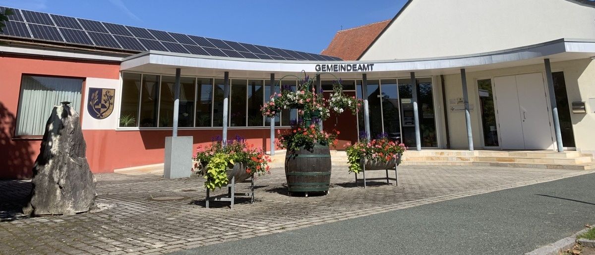 Ein Gemeindegebäude mit Solarpanelen auf dem Dach, einem roten Dach und einem grünen Turm. Vor dem Gebäude stehen einige Blumentöpfe.