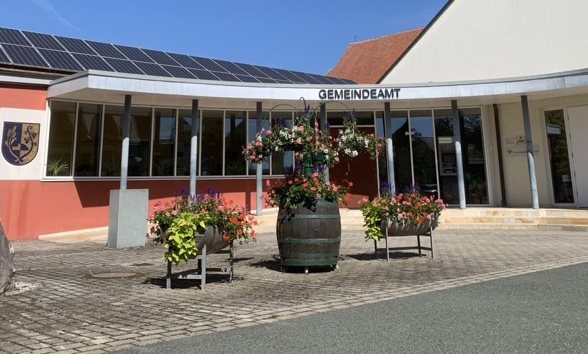 Ein Gemeindegebäude mit Solarpanelen auf dem Dach, einem roten Dach und einem grünen Turm. Vor dem Gebäude stehen einige Blumentöpfe.
