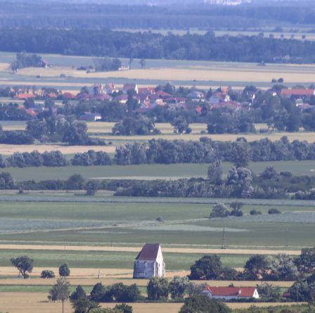Luftaufnahme einer ländlichen Landschaft mit Feldern, Häusern und Bäumen. In der Mitte sticht ein kleines Gebäude zwischen den grünen Feldern hervor. Am Horizont zeigen sich entfernte Hügel und ein klarer blauer Himmel.