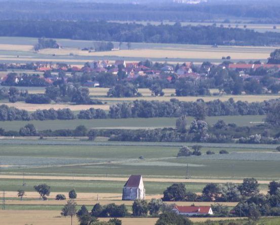 Luftaufnahme einer ländlichen Landschaft mit Feldern, Häusern und Bäumen. In der Mitte sticht ein kleines Gebäude zwischen den grünen Feldern hervor. Am Horizont zeigen sich entfernte Hügel und ein klarer blauer Himmel.