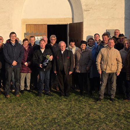 Eine Gruppe von Menschen posiert für ein Foto vor einem Gebäude mit einer Holztür. Einige tragen Brillen, Hüte und Schals. Einer hält eine Trophäe und Blumen.