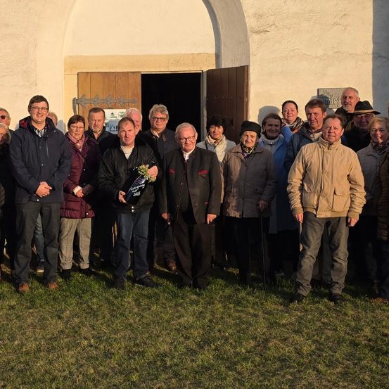 Eine Gruppe von Menschen posiert für ein Foto vor einem Gebäude mit einer Holztür. Einige tragen Brillen, Hüte und Schals. Einer hält eine Trophäe und Blumen.