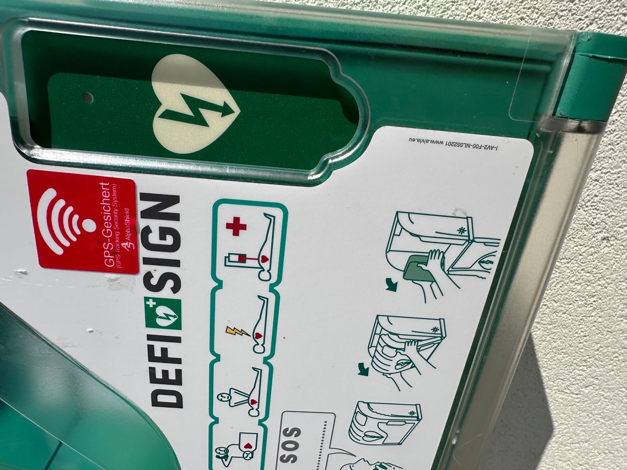 Eine grüne Defibrillatorbox mit dem Zeichen DEFI SIGN. Sie hat einen SOS-Notrufknopf und eine Anleitung zur Verwendung.