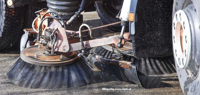 A street cleaning vehicle is parked on the side of the road. It has two large brushes and a rusty metal body.