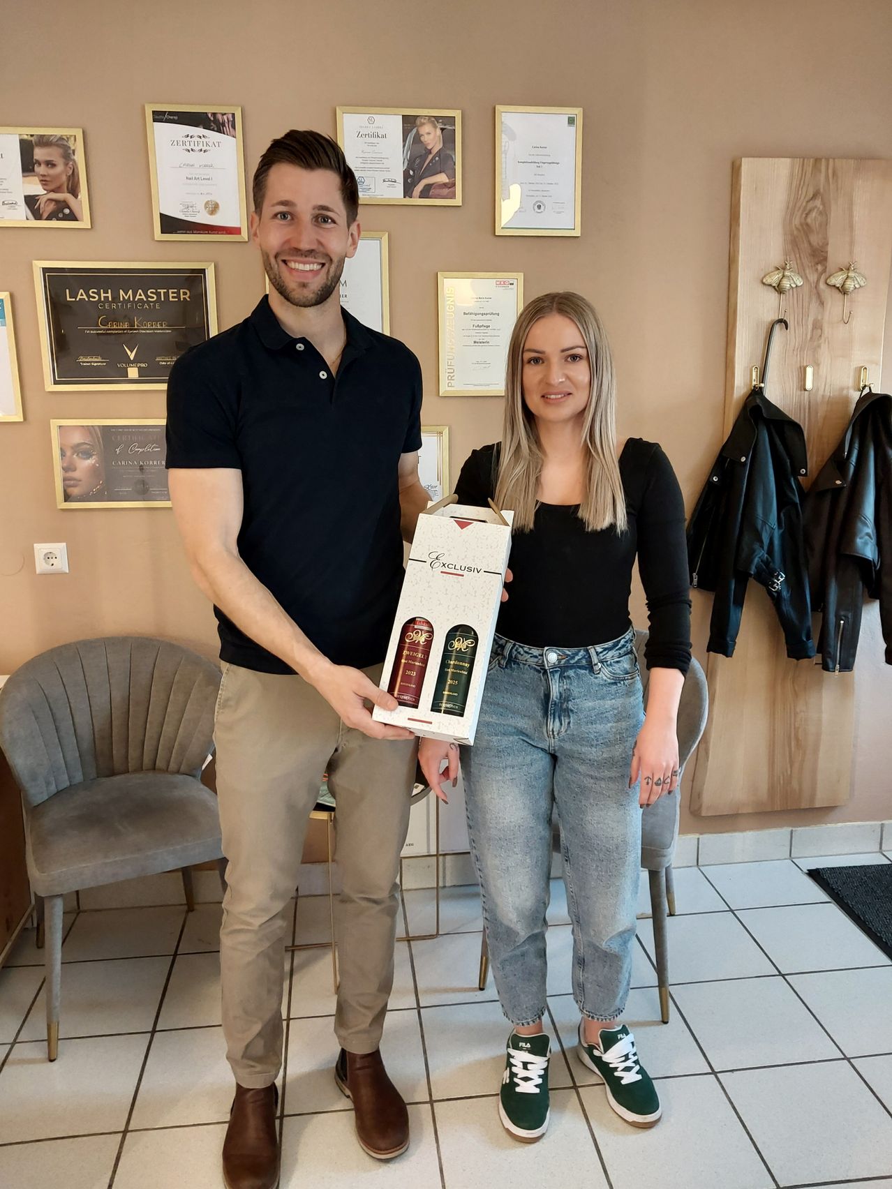 Ein Mann und eine Frau stehen lächelnd und halten eine Produktbox in einer Umgebung mit Zertifikaten an der Wand, hängenden Jacken und einem Stuhl im Hintergrund.