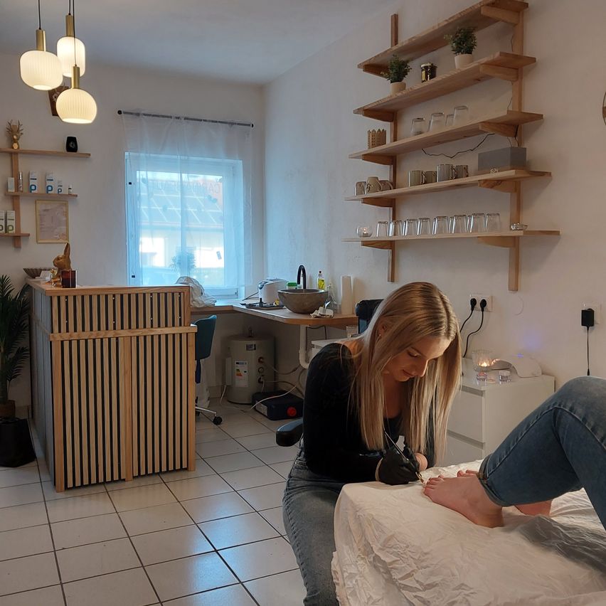 Eine Frau macht einer anderen Frau eine Pediküre in einem Salon, mit einem Schreibtisch, Regalen und einem Fenster im Hintergrund.