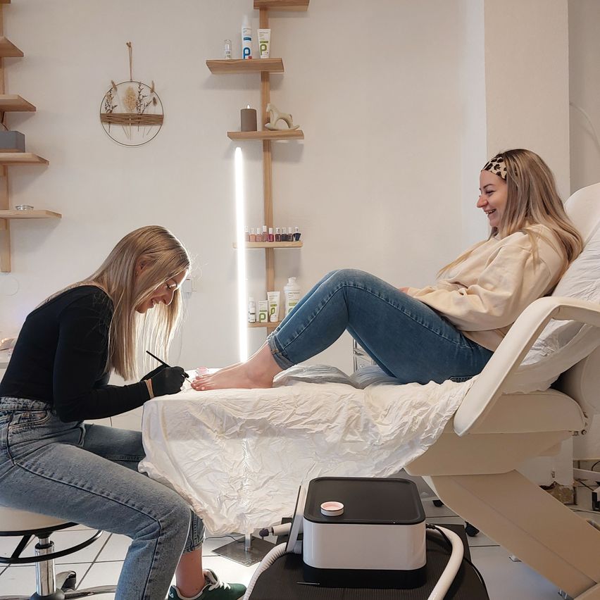 Eine Frau macht einer anderen Frau in einem Salon eine Pediküre, die auf einem Bett sitzt. Die Frau auf dem Bett lächelt.