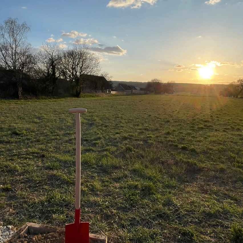 Ein leeres Feld mit einer Schaufel im Vordergrund bei Sonnenuntergang. Bäume und Häuser sind in der Ferne.