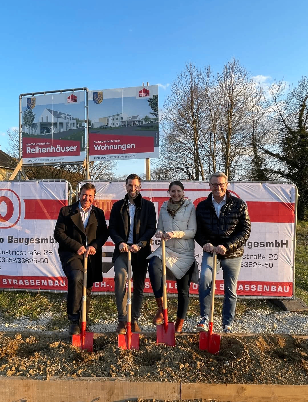 Vier Menschen posieren mit Schaufeln, wahrscheinlich bei einer Grundsteinlegungszeremonie, vor einem Banner mit 'Baugesmbh' und 'Reihenhäuser'. Dahinter befinden sich drei große Plakate, die neue Häuser bewerben.