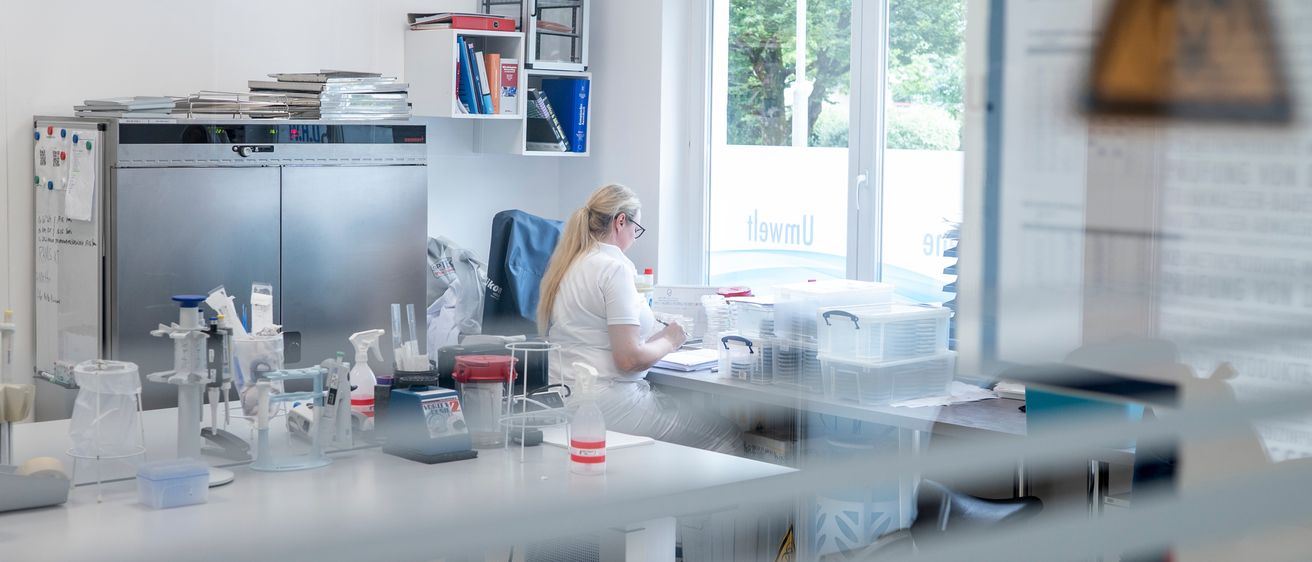 Eine Laborarbeiterin in Laborkittel und Brille arbeitet an einem Schreibtisch mit mehreren Behältern und Geräten. Das Labor ist gut beleuchtet, mit Regalen und einer Glastür hinter ihr. Ein Warnhinweis befindet sich an der Wand.