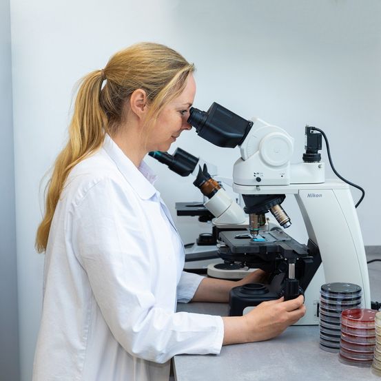 Eine Frau in einem weißen Laborkittel untersucht Proben mit einem Mikroskop in einem Labor.