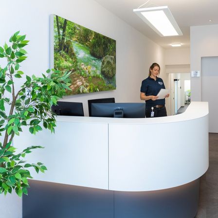 Eine Frau in einer blauen Uniform steht an einer Rezeptionstheke, hält eine Mappe und schaut in die Kamera. Hinter ihr ziert ein großes Gemälde die Wand, und zwei Computermonitore sind auf dem Schreibtisch.