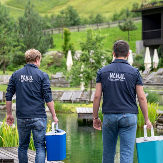 Zwei Männer in blauen Jacken mit W.H.U. auf dem Rücken, die Kühlboxen tragen, gehen auf ein Gewässer zu, mit einem Haus und Bäumen im Hintergrund.