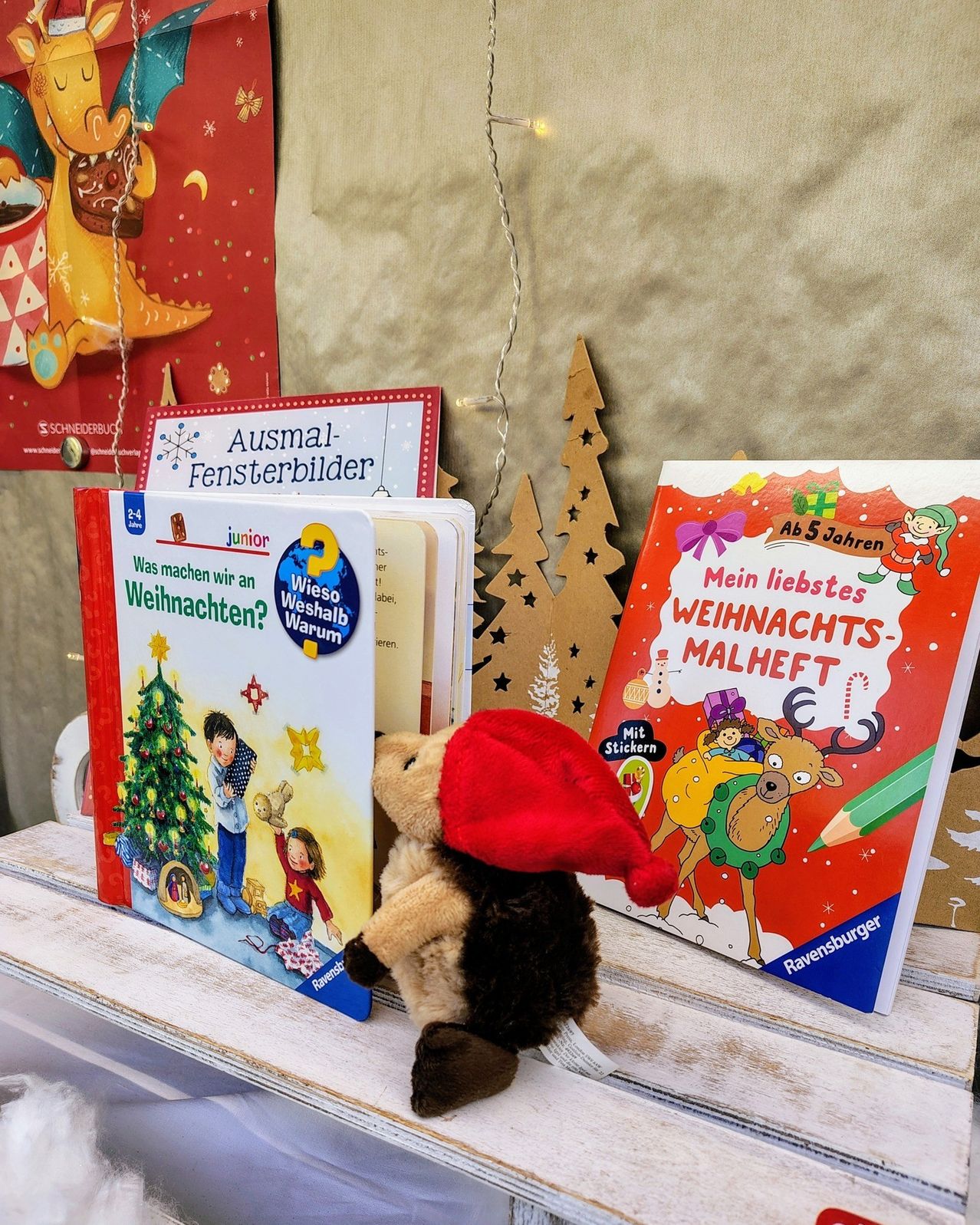 Ein roter Hut aus Stoff sitzt zwischen zwei Büchern auf einem weißen Regal. Die Bücher sind mit 'Was machen wir an Weihnachten?' und 'Mein liebstes Weihnachtsmalheft' betitelt.