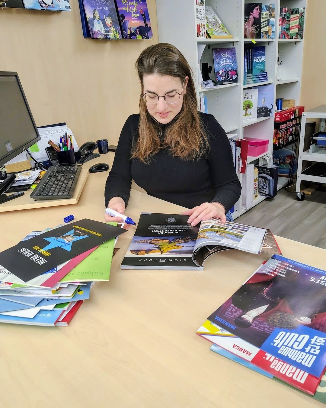 Eine Frau mit Brille sitzt an einem Schreibtisch mit Büchern, einem Computer und einem Marker und liest eine Zeitschrift.
