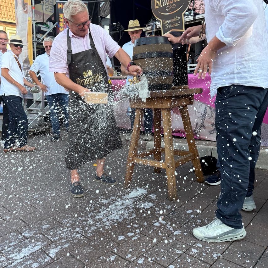 Ein Mann in einer schwarzen Schürze klopft mit einem Holzstab auf einen Holzfass, wodurch Bier auf einer Kopfsteinpflasterstraße spritzt.