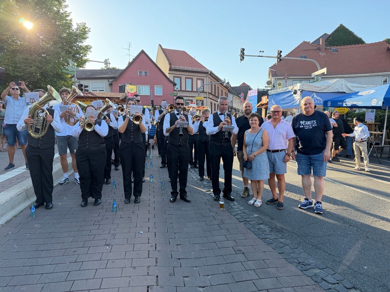 Eine Blaskapelle zieht die Straße entlang, vor ihnen steht eine Gruppe von Menschen. Die Bandmitglieder sind in formeller Kleidung gekleidet, und die Leute lächeln und posieren für ein Foto. Die Straße ist von Gebäuden gesäumt, und im Hintergrund befinden sich Straßenlaternen und Ampeln.