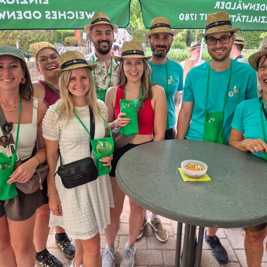 Eine Gruppe von Menschen in passenden grünen Lanyards und Hüten posiert für ein Foto im Garten. Ein Tisch mit einer Schüssel Nüssen ist im Vordergrund.