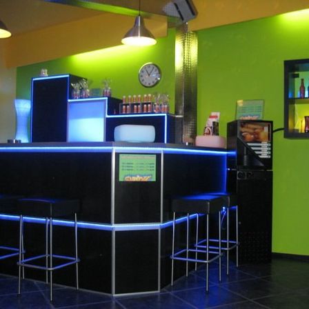 Eine Bar mit grünen Wänden und blauen Lichtern, mit Stühlen, einer Theke mit einem Schild, Regalen mit Flaschen, einer Uhr und Lampen.