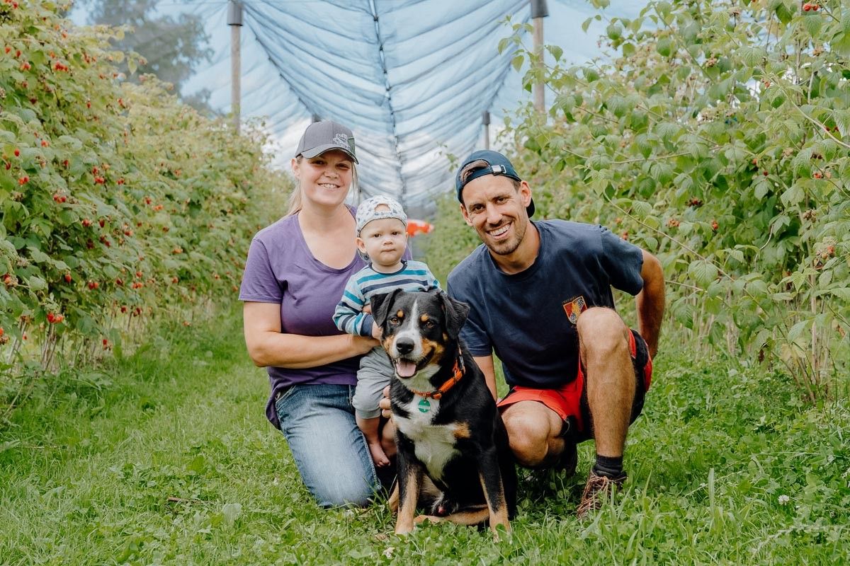 Eine Familie mit einem Baby, einem Hund und einem Mann in einer Kappe posiert für ein Foto in einem Beerenfeld.