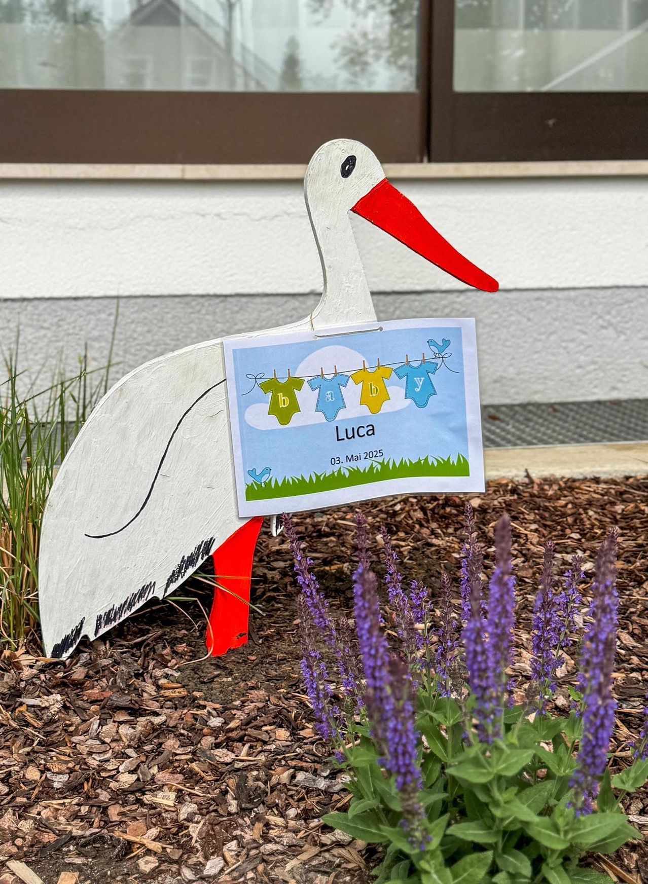 Eine Storchendekoration mit einer Karte, die Luca's Geburt am 3. Mai 2025 ankündigt. Die Karte ist am Storch befestigt und zeigt Babykleidung an einer Wäscheleine.