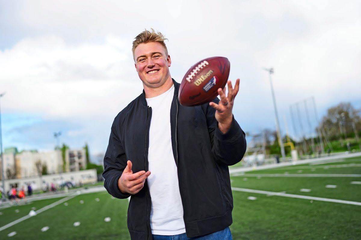 Ein Mann in einer schwarzen Jacke steht auf einem Footballfeld, lächelt und hält einen Football in den Händen.