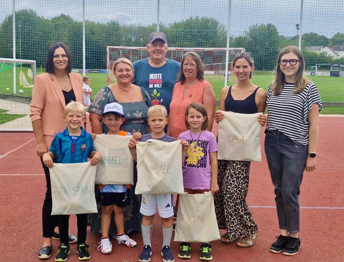 Eine Gruppe von Menschen auf einem Sportplatz, Erwachsene und Kinder, halten Taschen mit Namen. Sie scheinen für ein Foto zu posieren. Hinter ihnen steht ein Fußballtor, und in der Ferne sind Bäume und eine Straßenlaterne zu sehen.