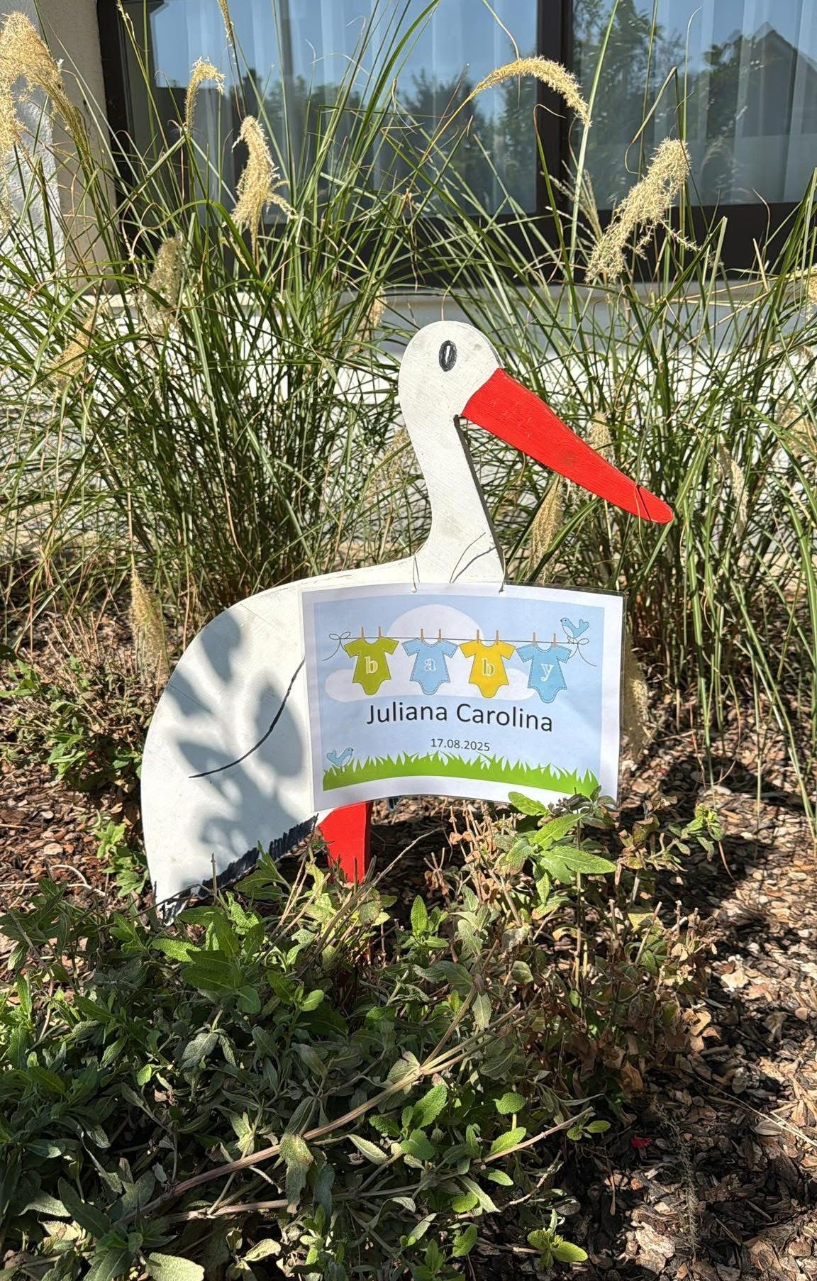 Eine Storchendekoration mit einem Schild, das Juliana Carolina und das Datum 17.08.2025 zeigt, in einem Garten mit hohem Gras platziert.