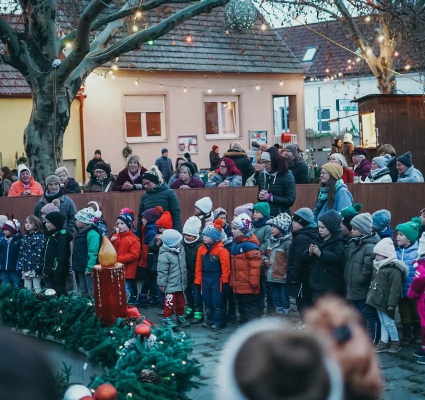 Eine Gruppe von Kindern und Erwachsenen versammelt sich um einen geschmückten Baum und eine brennende Kerze. Die Menge steht hinter einer Holzbarriere, mit Gebäuden und weiteren festlichen Lichtern im Hintergrund.