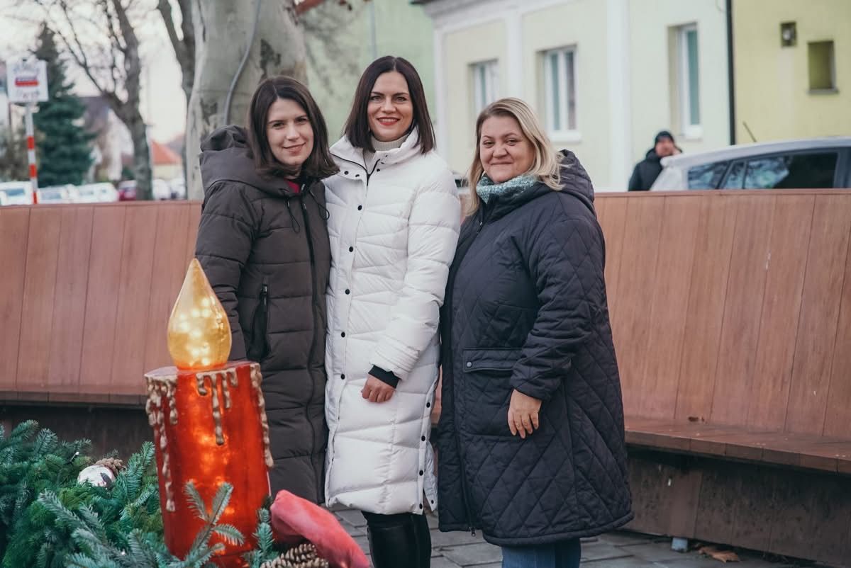 Drei Frauen in Wintermänteln stehen eng beieinander und lächeln für ein Foto. Eine hat einen weißen Mantel, eine einen dunkelbraunen und eine einen dunkelblauen. Sie stehen vor einer dekorativen Kerze und einem Holzzaun.