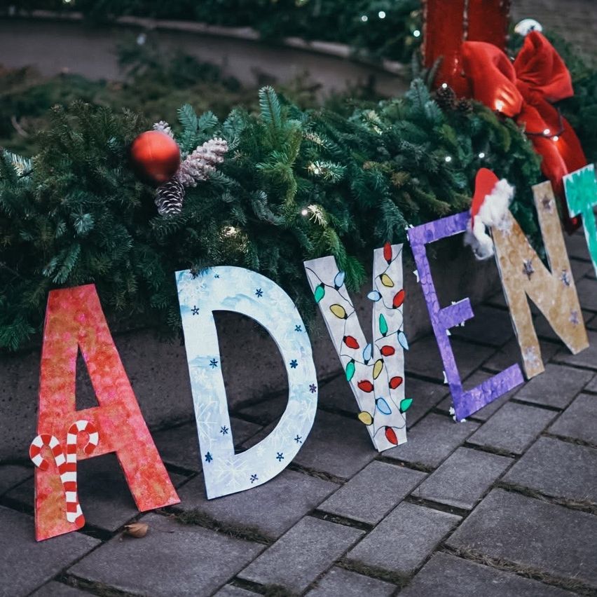 Das Wort 'ADVENT' ist in bunten Buchstaben auf einem Ziegelweg geschrieben, umgeben von festlichen Weihnachtsdekorationen.
