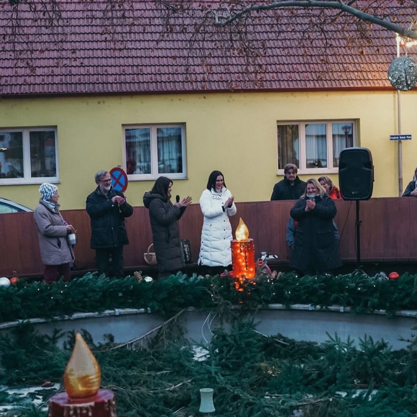 Eine Gruppe von Menschen steht um eine Kerze auf einer Plattform vor einem gelben Haus. Sie tragen Winterkleidung.